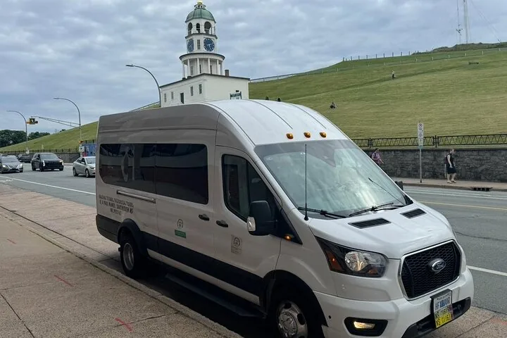 White tour van parked on a street near a grassy hill with clock tower